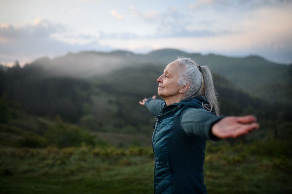 middle aged white woman with gray hair is seen enjoying nature as she hikes on a hillside at sunrise.